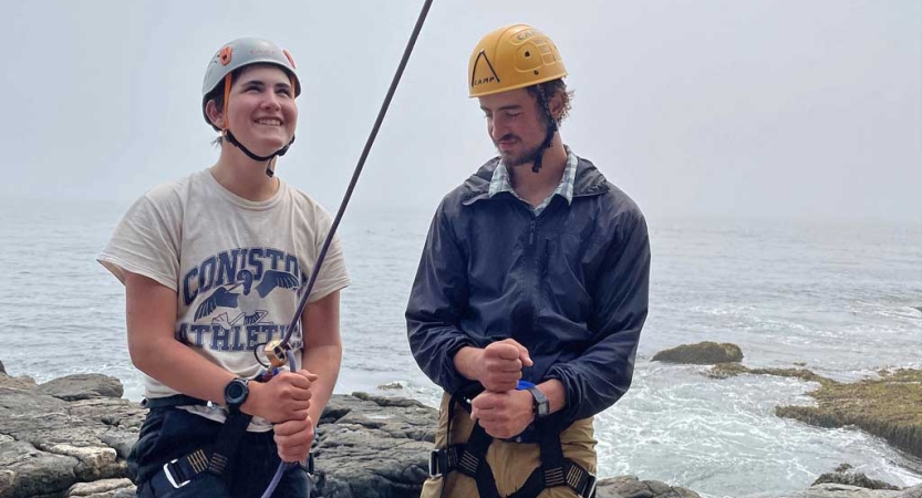 two people wearing safety gear stand on a rocky coast line and prepare to rock climb